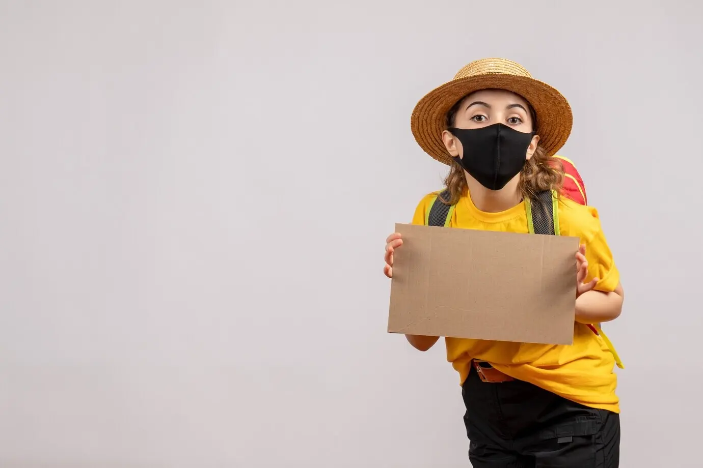 Vorderansicht einer weiblichen Reisenden mit Rucksack, die vor einer grauen Wand ein Stück Pappe hochhält.